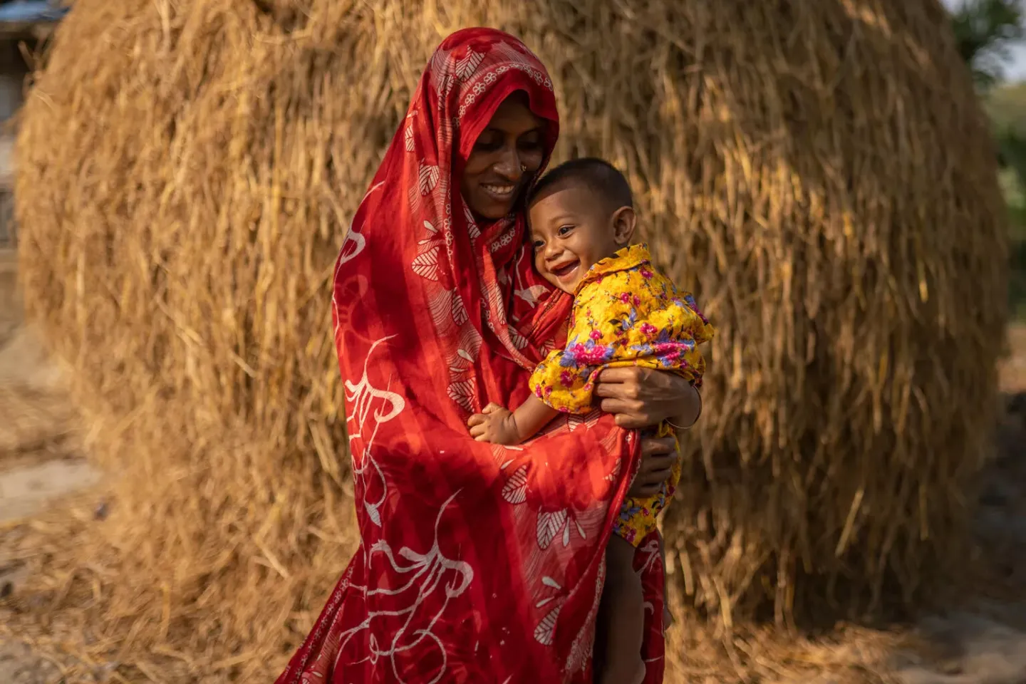 A woman wearing a bright red patterned garment holds a young child against her body. The child is wearing yellow/gold patterned clothing. They are standing outdoors in front of dried grass stacked in a shelter. 